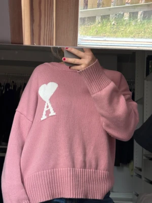 Rosa stickad tröja från Ami - INTREKOLL!!! Superfin rosa stickad tröja från Ami med ett vitt hjärta och bokstaven A på bröstet. Tröjan har en loose passform, ribbade muddar och rund hals. Perfekt för dig som gillar snygga detaljer och vill ha en mysig vibe. Såå fin och cool🩷🩷🩷