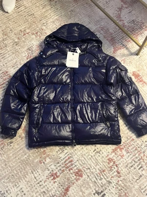 Mörkblå Moncler pufferjacka med huva - Säljer en mörkblå pufferjacka från Moncler med glansig finish och huva. Jackan har dragkedja framtill, två sidofickor med dragkedja och klassisk Moncler-logga på ärmen. Insidan har ett coolt serietidningsmotiv. Perfekt för kalla dagar!