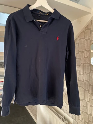 Marinblå långärmad pikétröja Polo Ralph Lauren - Säljer en marinblå långärmad pikétröja från Polo Ralph Lauren i slim fit. Tröjan har klassisk krage, knäppning vid halsen och den ikoniska röda loggan broderad på bröstet. Liten i storleken så passar nog S/M