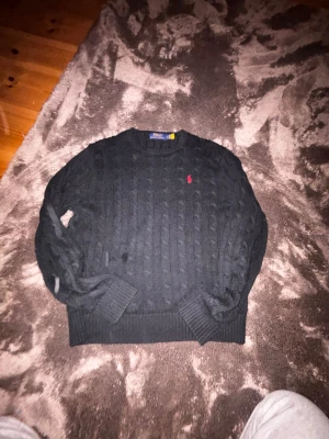 Svart kabelstickad tröja Polo Ralph Lauren - Svart kabelstickad tröja från Polo Ralph Lauren i storlek L. Klassisk design med rund hals och röd broderad logga på bröstet. Tröjan har ribbade muddar vid ärmslut och nederkant, perfekt för dig som gillar stilren och tidlös look.