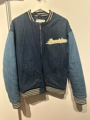 ASOS Design jeans bomberjacka - Säljer en jeans bomberjacka från ASOS Design med broderade detaljer. Mörkblå kropp och ljusare blå ärmar, broderad text 'Brooklyn' fram och 'New York' bak. Randiga muddar och krage, dragkedja framtill. Obs: Alla plagg säljs endast fram till 28/12