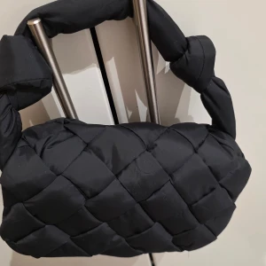 Svart quiltad handväska Carin Wester - Quiltad svart handväska från Carin Wester, modell Marie Padded Bag. Väskan har en flätad axelrem och är tillverkad i 100% polyester. Magnetknäppning och flera fack inuti, inklusive ett med dragkedja. Perfekt storlek för det viktigaste och riktigt snygg design.