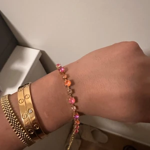 Guldarmband med rosa och orange stenar - Snyggt guldarmband från Caroline Svedbom med glittriga stenar i rosa och orange nyanser. Armbandet har justerbar kedja och en liten hjärtformad bricka med varumärket ingraverat. Perfekt för dig som gillar färg och bling!