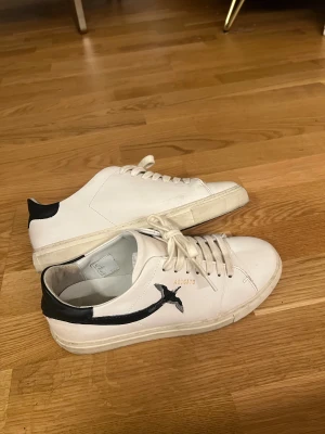 Vit sneaker från Axel Arigato - Snygga vita sneakers från Axel Arigato med svart detalj på hälen och en broderad svart fågel på sidan. Skorna har snörning och en klassisk, rund tå. Tillverkade i skinn och har en stilren, minimalistisk design med guldfärgad logga.