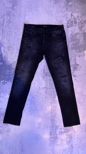 Svarta slim jeans med slitningar  - Säljer ett par snygga svarta slim fit jeans från Jack & Jones i storlek (31-30) med snygga slitningar och distressed detaljer på benen. Jeansen har klassisk femficksmodell. 