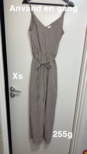 Rutig byxdress med knytband XS - Säljer en snygg rutig byxdress i beige och svart med smala axelband och knytband i midjan. Lätt och luftig modell med raka ben, perfekt för varma dagar. Passar dig som gillar en avslappnad men trendig stil. 🌸 Använd en gång. Storlek Xs