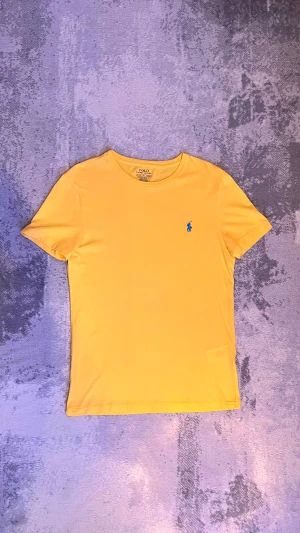 Gul t-shirt från Polo Ralph Lauren - Snygg gul t-shirt från Polo Ralph Lauren med klassisk blå logga broderad på bröstet. T-shirten har rund hals och korta ärmar, perfekt för dig som gillar stilrena och färgglada plagg. Materialet är mjuk bomull som känns skönt mot huden.