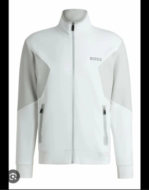limited edition Vit trackjacka från BOSS  - Snygg vit trackjacka från BOSS med ljusgrå paneler på ärmar och sidor. Jackan har hög krage, hel dragkedja framtill och två fickor med dragkedja. Materialet är mjukt och känns sportigt, perfekt för chill eller träning. BOSS-logga på bröstet ger en clean look. PRIS KAN DISKUTERAS!!