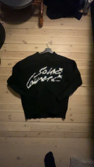 Svart stickad Saint Laurent tröja - Säljer en svart stickad tröja från Saint Laurent med vit, stor logga i graffiti-stil över bröstet. Tröjan har lång ärm och en loose passform, perfekt för dig som vill ha en cool och trendig look. Passar kring xs.                                               Byten kan vara intressant!