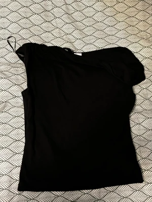 Svart oneshoulder topp från DIVIDED - Säljer en svart oneshoulder topp från DIVIDED i storlek XXS. Toppen har en asymmetrisk design med en axelbandslös sida och ett smalt axelband på andra sidan. Materialet är mjukt och stretchigt, perfekt för att skapa en cool och trendig look.