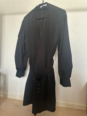 Svart omlottklänning från NA-KD - Svart omlottklänning från NA-KD breda långa ärmar/ballongärmar. Klänningen har ett bälte med stort spänne i brunt och silver, samt v-ringning.  Obs: Alla plagg säljs endast fram till 28/12 