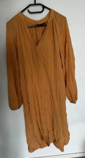 Klänning från KappAhl - Säljer en orange klänning från KappAhl i storlek M. Klänningen har en avslappnad passform med långa ärmar och knappar framtill. Materialet är mjukt och krinklat.  Obs: Alla plagg säljs endast fram till 28/12 
