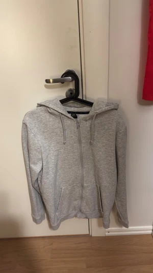 zip-hoodie  - Snygg grå hoodie från SOC i storlek S. Den har dragkedja hela vägen, två fickor fram och en skön huva med dragsnören. Mjuk och bekväm bomullsblandning, perfekt för chill eller träning. Enkel och stilren design som funkar till det mesta.
