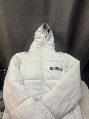 Vit pufferjacka från Hoodrich  - Säljer en clean vit pufferjacka från Hoodrich i storlek M. Jackan har broderad logga på bröstet och tryckt logga på insidan av kragen, samt dragkedja framtill. Fylld med syntetmaterial och har en hög krage för extra värme. Perfekt för kalla dagar. Litet sltiage under luvan, se sista bilden, annars fint skick, något ljus brun i inner ärmarna! Nypris = 1500