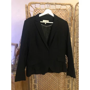 Svart kavaj från KappAhl, storlek 42 - Stilren svart kavaj från KappAhl i klassisk modell med smal krage och knappstängning framtill. Kavajen har två fickor med lock och är helfodrad för en snygg siluett. Perfekt att matcha med jeans eller kostymbyxor för en cool look. Använd en gång.