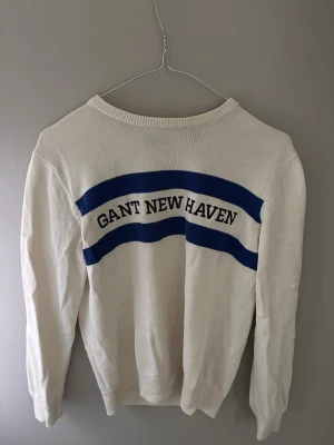 Vit stickad tröja GANT New Haven - Vit stickad tröja från GANT med blåa ränder och texten 'GANT NEW HAVEN' på ryggen. Tröjan har rund hals och långa ärmar, perfekt för dig som gillar klassisk och sportig stil. Materialet är mjukt och skönt bomull.