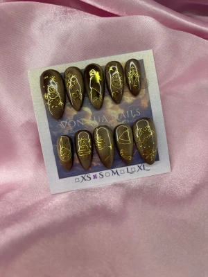 CocoaXmas (S);Medium Almond - Set bruna cateye press-on naglar i storlek S: 15 mm, 11,5 mm, 12 mm, 11 mm, 9 mm , dekorerade med guldiga julmotiv som snögubbe, julgran, julkula och texten 'Merry Christmas'. Naglarna har en medium mandelformad design och skimrande cateye finish, perfekta för dig som vill ha extra festliga naglar under vintern.