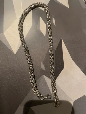 Tungt silverfärgat kedjehalsband - Säljer ett robust halsband i silverfärgad metall med ett coolt, flätat kedjemönster. Låset är av karbinmodell och hela halsbandet har en chunky vibe som sticker ut. Perfekt för dig som gillar statement-smycken och vill ha något som syns.