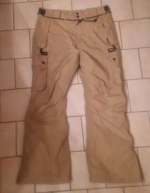 Beiga täckbyxor från Dope Snow L - Säljer ett par beiga täckbyxor från Dope Snow i storlek L. Byxorna har flera praktiska fickor med lock och knappar, snörning i midjan och dragkedjor vid bensluten. Perfekta för vintern och outdoor-aktiviteter. Materialet är slitstarkt och vattenavvisande. Några små revor på vänsterbenet, syns knappt