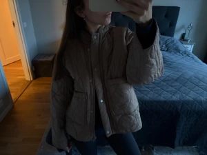 Beige quiltad pufferjacka med fickor - Säljer en beige pufferjacka med quiltad design och stora framfickor. Jackan har hög krage, dragkedja och knappar framtill. Modellen är loose fit och har långa ärmar. Perfekt för dig som gillar en chill och trendig look.