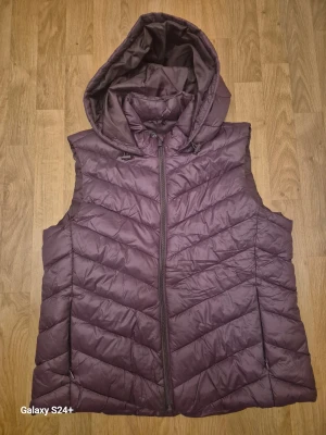 Mörklila pufferväst från Primark - Säljer en mörklila pufferväst från Primark med huva och dragkedja framtill. Västen har ett quiltat mönster och är tillverkad i syntetmaterial. Perfekt för lager-på-lager och chill höst- eller vinterstil.