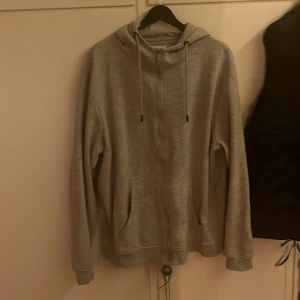 Grå hoodie från Denim Day XL - Grå hoodie från Denim Day i storlek XL. Tröjan har dragkedja framtill, huva med snörning och två fickor. Materialet är mjukt och känns som bomull, perfekt för chill dagar. Klassisk passform med långa ärmar och ribbade muddar.