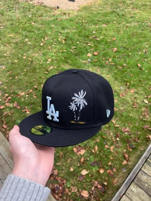 Svart LA Dodgers 59FIFTY keps - Snygg svart 59FIFTY keps från New Era med LA Dodgers-logga i vitt framtill, broderade palmer och en patch med Dodger Stadium på sidan. Kepsen har även ett litet broderat hjärta bak. Klassisk flat brim och cool streetstil.