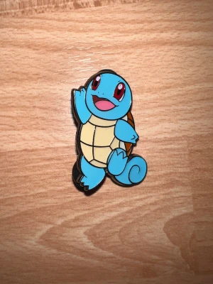 Pokémon Squirtle pin i metall - Säljer en officiell Pokémon pin med Squirtle-motiv. Pin:en är gjord i metall med färgglad emalj i blått, beige och brunt. Den har två svarta gummibakstycken för säker fastsättning. Perfekt för jackan, väskan eller samlingen!