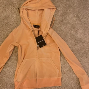 Persikofärgad Juicy Couture hoodie XS - Juicy Couture hoodie i mjuk persikofärgad velour med dragkedja och huva. Klassisk JC-logga broderad på bröstet och stora fickor framtill. Perfekt för en chill och trendig look.