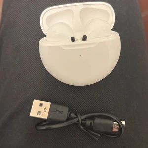 Trådlösa hörlurar med laddningsladd. - Snygga vita trådlösa hörlurar med tillhörande laddningsetui och USB-laddningskabel. Hörlurarna är i gott skick utan synligt slitage och fungerar utmärkt för musik. Perfekt för dig som vill ha frihet och bra ljudkvalitet på språng.