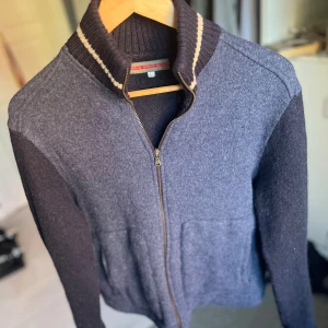 Mörkblå stickad cardigan från Bray Steve Alan - Snygg mörkblå stickad cardigan från Bray Steve Alan med ribbad krage och vita ränder. Jackan har dragkedja framtill och är tillverkad i ull, vilket gör den både mjuk och varm. Klassisk design med långärmade ärmar och stilren look, perfekt för dig som gillar enkel men cool stil. Den är lite kortare i i längden av tröjan alltså vid midjan men är inget märkbart. Den passar storlek M mer än L. Hör av dig vid minsta fråga🙌