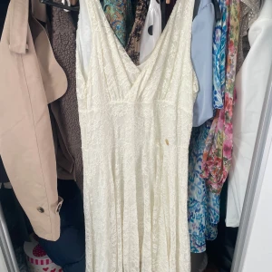 Vit lång spetsklänning omlott - Romantisk långklänning i vit spets med omlott och djup v-ringning. Klänningen har ett luftigt fall och är perfekt för sommarens alla tillfällen. Spetsdetaljerna ger en bohemisk och elegant känsla.