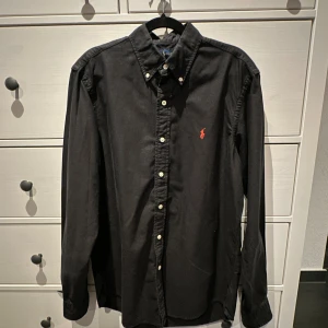 Svart skjorta från Ralph Lauren - Klassisk svart skjorta från Ralph Lauren med röd broderad logga på bröstet. Skjortan har knappar hela vägen, button-down krage och långa ärmar. Perfekt för en stilren look.
