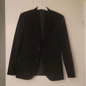 Svart kavaj Skinny Fit från H&M - Snygg svart kavaj från H&M i modellen Skinny Fit. Klassisk design med två knappar framtill och smal passform. Perfekt för dig som gillar en stilren och modern look. Fodrad insida för extra komfort.