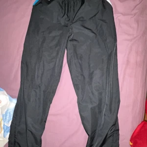 Svarta träningsbyxor Lacoste Sport M - Svarta träningsbyxor från Lacoste Sport i storlek M. Byxorna har elastisk midja, blå detalj på sidan och klassiska Lacoste-loggan framtill. Lätta och bekväma, perfekta för sport eller chill. Materialet är syntet och andas bra.