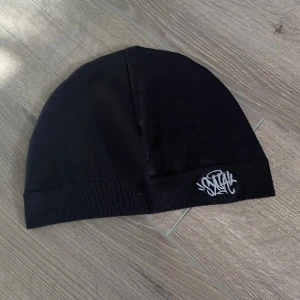 Svart Syna world mössa - Svart, tight mössa från syna world med en broderad vit logga framtill. Mössan har en enkel och stilren design med ribbad kant nertill. Perfekt för dig som gillar sportig och clean stil. Materialet känns mjukt och stretchigt, troligen polyester. Köpte in fel matrial det skulle vara annorlunda denna blev mer som gummi men funkar fortfarande att ha på och se snyggt ut