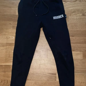 Svarta mjukisbyxor från Hoodrich - Svarta mjukisbyxor från Hoodrich med vit logga på ena benet. Byxorna har snörning i midjan och smal passform nedtill. Perfekta för en avslappnad och sportig stil.
