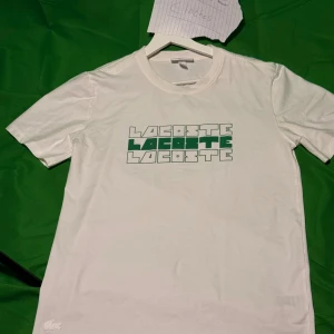 Vit Lacoste t-shirt med grön logga - Vit t-shirt från Lacoste i slim fit med coolt tryck av Lacoste-loggan i grönt och vitt på bröstet. Klassisk rund hals och korta ärmar. Tillverkad i mjuk bomull för skön känsla. Perfekt för en clean och stilren look.Liknande modell som originalet!