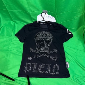 Svart t-shirt med döskalle från Philipp Plein - Svart t-shirt från Philipp Plein med glittrande döskalle och korslagda ben på framsidan samt stor text 'PLEIN' i stenar. Kortärmad modell med logotypdetalj på ärmen och etikett i sidan. Perfekt för dig som gillar edgy och lyxig streetstyle.Liknande modell som originalet!