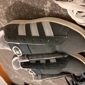 Adidas Campus grå sneakers - Säljer ett par Adidas Campus sneakers i mörkgrå mocka med vita detaljer och klassiska tre ränder på sidorna. Skorna har vit sula, rund tå och kommer med både grå och vita skosnören. Perfekta för dig som gillar retrostil och streetwear.