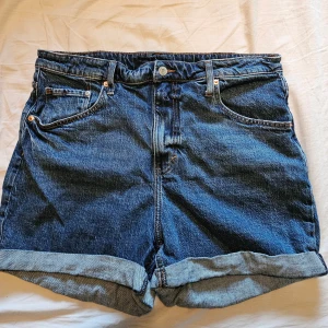 Denim shorts - Säljer ett par klassiska blå mom shorts från H&M med hög midja och regular fit. De har uppvikta benslut, fem fickor och knapp- samt dragkedjestängning. Aldrig använda endast tvättade