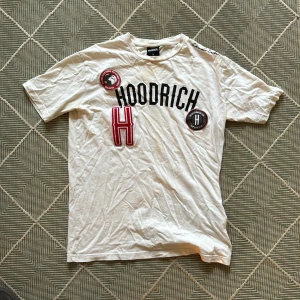 Vit Hoodrich t-shirt med tryck - Vit t-shirt från Hoodrich med svart logotyptryck och stora röda och svarta detaljer framtill. Klassisk passform med rund hals och korta ärmar. Skön bomullskvalitet och cool streetstil, perfekt för dig som gillar statement-plagg.