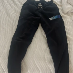 Svarta mjukisbyxor från Hoodrich XS - Svarta mjukisbyxor från Hoodrich i storlek XS med broderad logga i vitt och blått på ena benet. Byxorna har resår och snörning i midjan för skön passform. Perfekta för chill eller streetwear-stil.