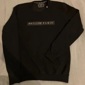 Svart Philipp Plein sweatshirt S - Svart sweatshirt från Philipp Plein med logga framtill och diskret metallmärke vid nederkanten. Klassisk rund halsringning och långa ärmar. Mjuk och skön bomullskvalitet, perfekt för en clean och stilren look till skolan eller att ha till fritiden.
