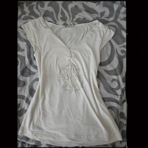 2000s top  - Säljer en vit blus med v-ringning och korta ärmar. Toppen har ett diskret broderat mönster framtill som ger en cool detalj. Materialet känns mjukt och lätt, perfekt för varmare dagar. Passformen är normal och toppen är enkel att matcha med jeans eller kjol.