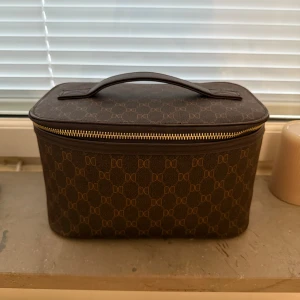 Brun handväska från Don Donna - Snygg brun handväska från Don Donna med klassiskt monogrammönster och guldfärgad dragkedja. Väskan har en rektangulär form, praktiskt handtag upptill och är tillverkad i syntetmaterial. Perfekt för dig som gillar stilrena accessoarer med logga.