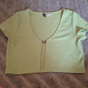 Grön croppad topp från H&M Divided - Snygg ljusgrön croppad topp från H&M Divided i ribbad bomullsmix. Toppen har korta ärmar, djup v-ringning och en söt fjärilsdetalj framtill som stängning. Perfekt för att ge din outfit en fräsch och trendig vibe.