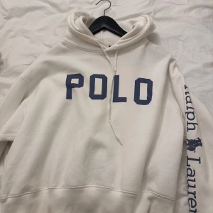 Vit hoodie från Polo Ralph Lauren - Vit hoodie från Polo Ralph Lauren med blått tryck 'POLO' på bröstet och 'Ralph Lauren' längs ena ärmen. Klassisk huva med snörning och avslappnad passform. Perfekt för en chill och stilren look.
