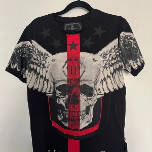 Svart t-shirt med dödskalle från Philipp Plein - Svart t-shirt från Philipp Plein med stort tryck av en dödskalle med vingar och röda detaljer framtill. T-shirten har korta ärmar och är gjord i mjuk bomull. Snygga stjärnor upptill och PP-logga i nacken. Perfekt för dig som gillar edgy streetstyle.