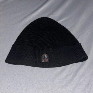 Svart ribbad mössa från Parajumpers - Svart ribbstickad mössa från Parajumpers med klassisk uppvikt kant och broderad PJS-logga framtill. Tillverkad i mjuk ullblandning som håller dig varm. Perfekt accessoar för kyliga dagar och snygg till streetwear. Har fått den i present 2024 Vintras använd 2-3 gånger 
